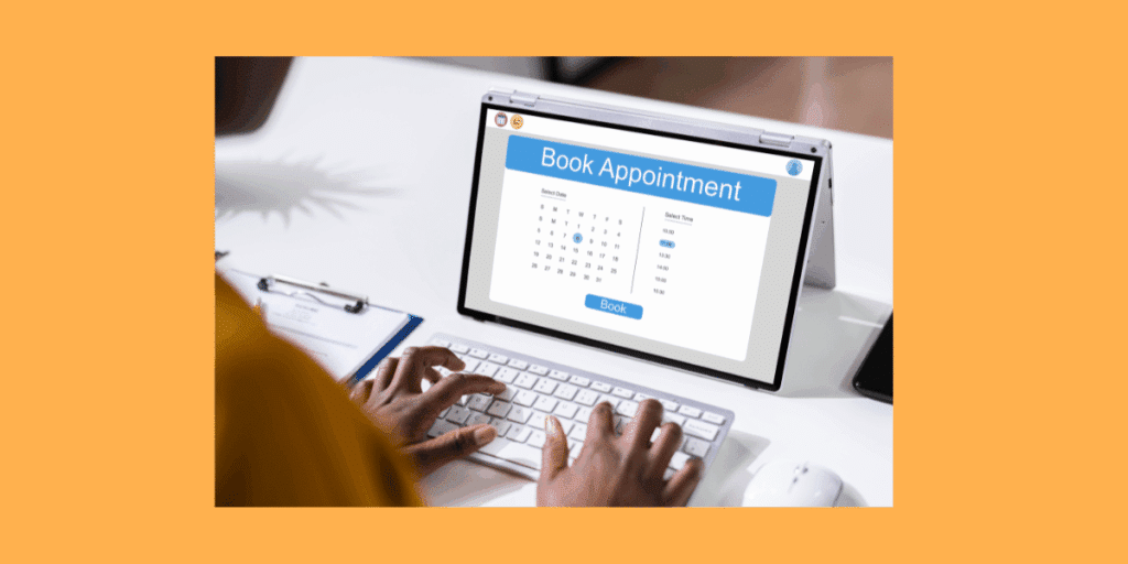 A person with an orange shirt is using a wireless keyboard to interact with a silver laptop. The laptop screen displays a web page for booking a doctor's appointment, showing a monthly calendar grid."