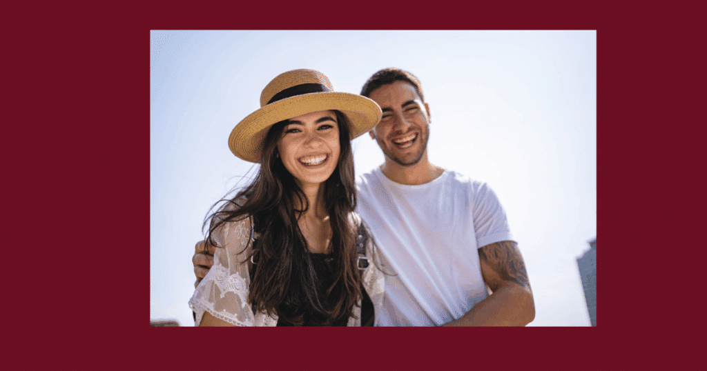 A smiling young couple standing close together outdoors; the woman wears a wide-brimmed straw hat, while the man has his arm around her, displayed on a burgundy background.